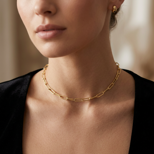 A close-up lifestyle shot of a model wearing a delicate gold paperclip chain necklace and a matching small gold stud earring, styled with a black velvet top against a warm, softly blurred background.