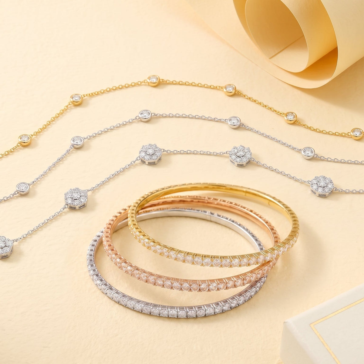 A jewelry flat-lay featuring delicate tennis-style necklaces in gold and silver with round clear stones, alongside three matching bangles in yellow, rose, and silver gold tones. The items are arranged on a warm beige background with a rolled cream paper and a small box in the background.