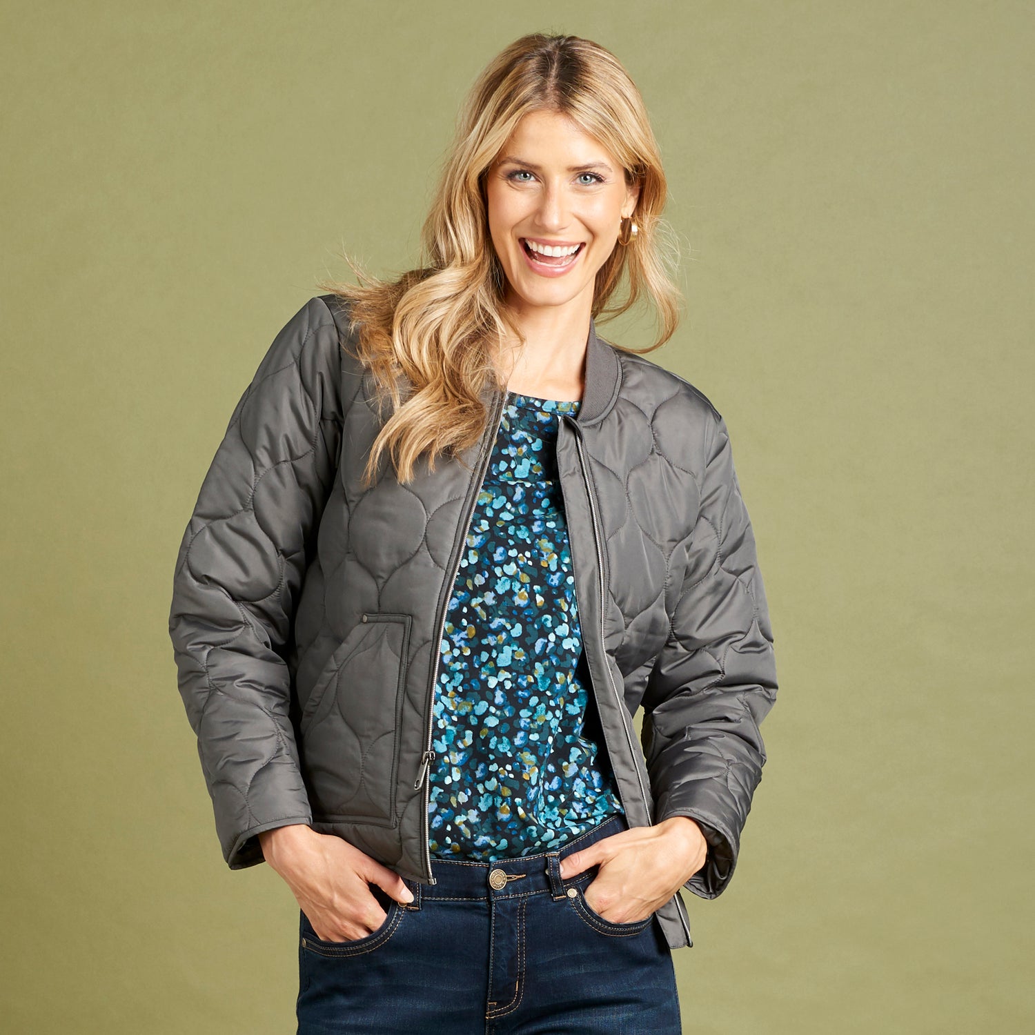 Woman wearing a gray quilted jacket, blue floral top, and dark jeans on a green background