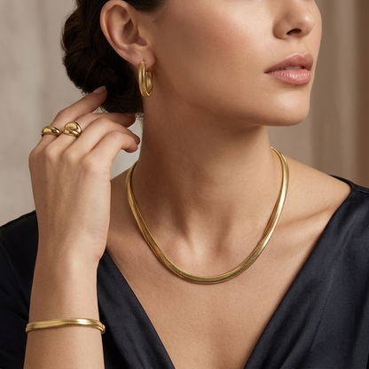 A close-up lifestyle shot of a model wearing a gold snake chain necklace, matching gold hoop earrings, and a gold bangle bracelet, paired with a dark navy satin top.